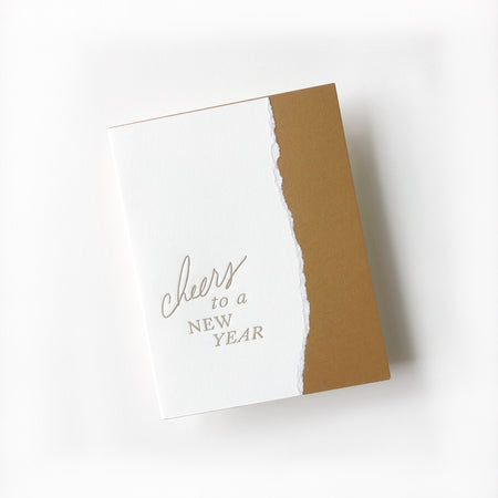 White and gold card with curved vertical line where colors meet and gold text saying, “Cheers to a New Year”. A white envelope is included.