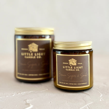 Brown glass jar with gold lid and brown label with gold foil text saying, “Little Light Candle Co. Brown Sugar & Smoke”. 