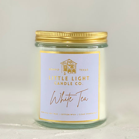 Glass jar with gold lid and lilac label with gold foil text saying, “Little Light Candle Co. White Tea”.