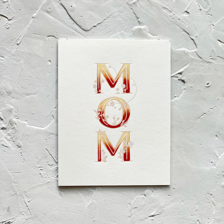 Ivory card with variegated colored text in orange, red and white saying, “MOM”. Images of embossed flowers. A gray envelope is included.