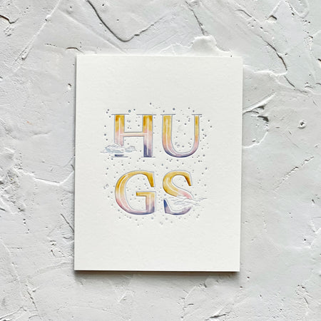 Ivory card with variegated text in gold, white, pink and blue saying, “HUGS”. Images of clouds and embossed circles. A gray envelope is included.
