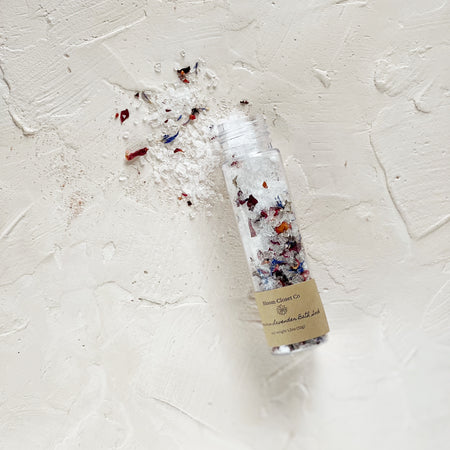 Small glass bottle with brown label saying, “Bloom Closet Co. Luxurious Lavender Bath Salts”. White salt mixed with brown, black and blue flecks of color.