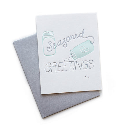 White card with gray and teal text saying, “Seasoned Greetings”. Images of teal salt and pepper shakers. A gray envelope is included.