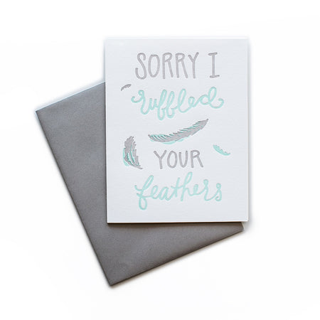 White card with gray and teal text saying, “Sorry I Ruffled Your Feathers”. Images of gray and teal feathers. A gray envelope is included.