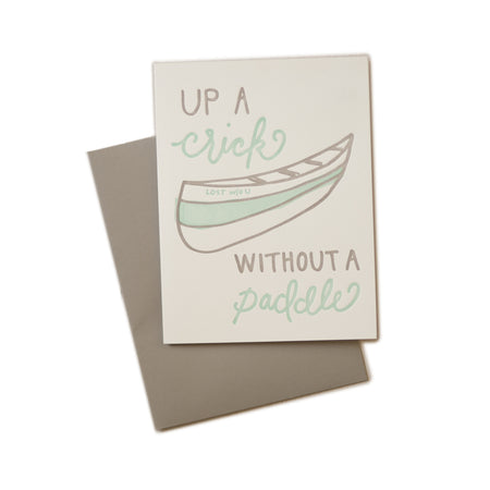 White card with gray and teal text saying, “Up A Crick Without A Paddle”. Image of a gray and teal canoe. A gray envelope is included.