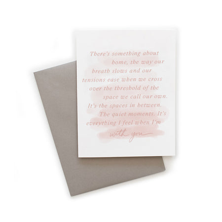 White card with watercolor wash background in pink tones with pink text saying, “There’s something about home, the way out breath slows and our tensions ease when we cross over the threshold of the space we call our own. It’s the spaces in between. The quiet moments. It’s everything I feel when I’m with you”. A gray envelope is included.