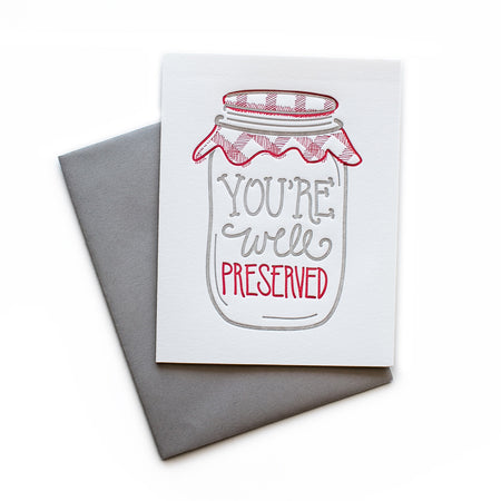 White card with gray and red text saying, “You’re Well Preserved”. Image of a gray and red jar with red checkered lid. A gray envelope is included.