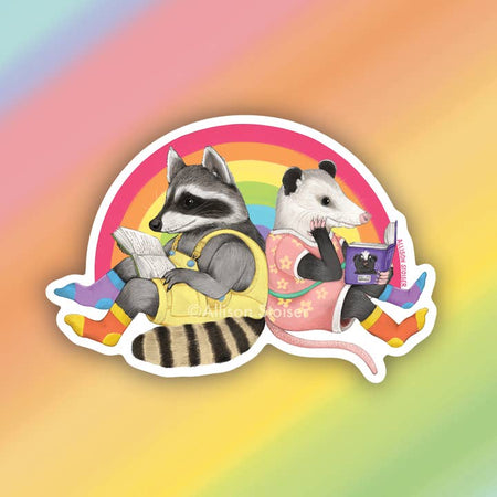 Sticker in the image of a raccoon wearing yellow overalls and multicolored socks and an opossum wearing a pink dress with flowers and multicolored socks. Raccoon and opossum are sitting back to back reading books.