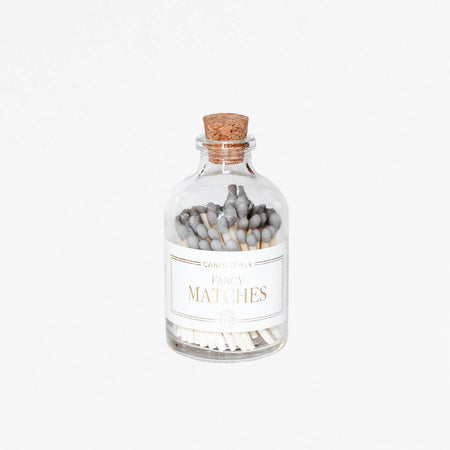 Small glass bottle with cork lid and white label with gold text saying, “Candlefolk Fancy Matches”. Filled with wooden matches with grey tops.