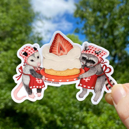 Sticker in the image of a raccoon and an opossum dress in red and pink chef hats and aprons holding a piece of strawberry shortcake on a red plate.