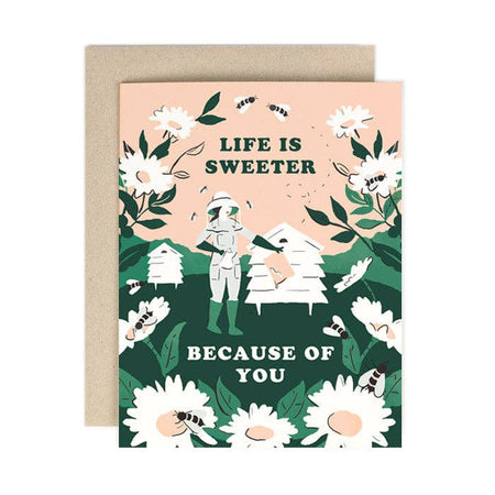 Ivory card with green and white text saying, “Life is Sweeter Because of You”. Images of a woman beekeeper with a bee hive surrounded by white daisies and bees. A brown envelope is included.