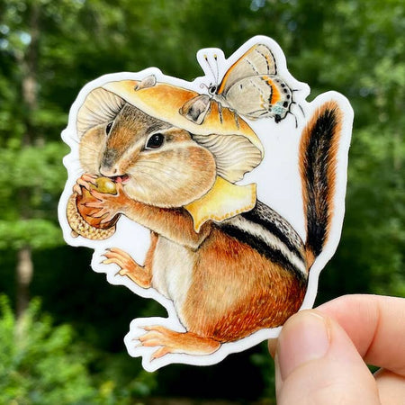 Sticker in the image of a chipmunk wearing a mushroom cap hat and eating an acorn.