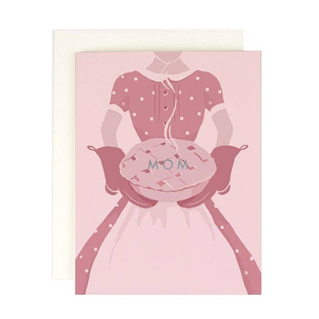 Pink card with image of a mother wearing a pink dress and apron with pink oven mitts holding a warm pie. Silver foil text across pie saying, “MOM”. An ivory envelope is included.