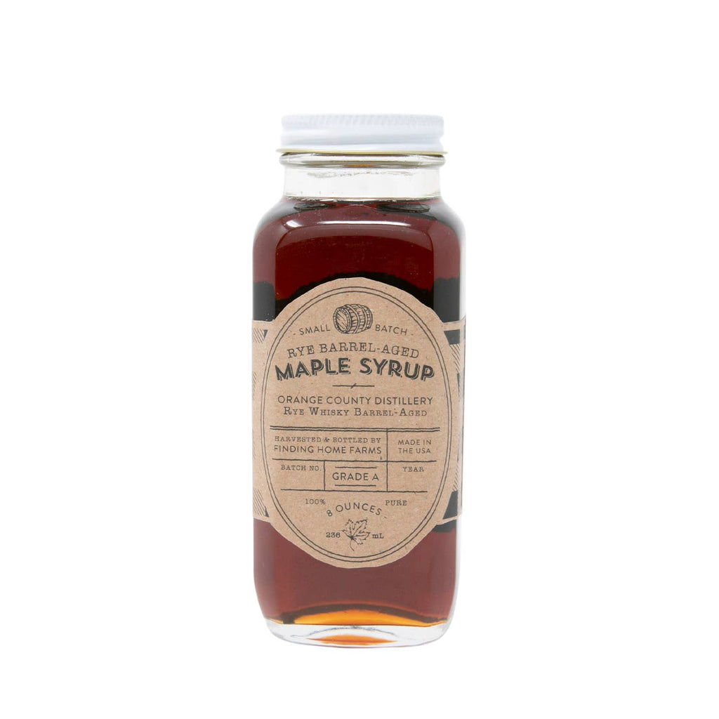 Glass bottle with white lid and brown label with black gingham checkered border. Black text saying, “Finding Home Farms Rye Barrel Aged Maple Syrup”.