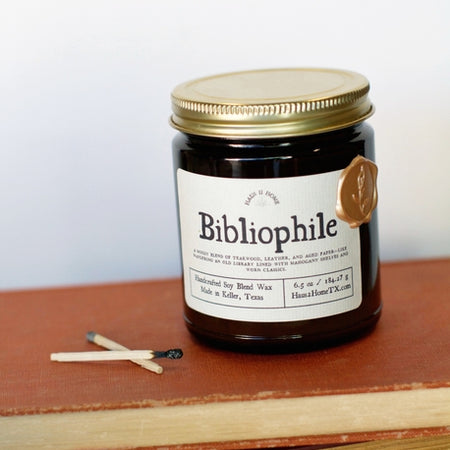 Amber color jar candle with gold lid. White label with black text saying, “Bibliophile”.