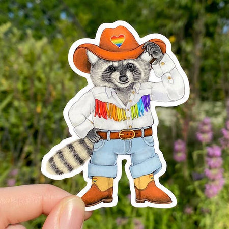 Sticker in the image of a raccoon wearing a brown cowboy hat with a rainbow heart in the center, a white shirt with rainbow colored fringe, blue jean and brown cowboy boots.