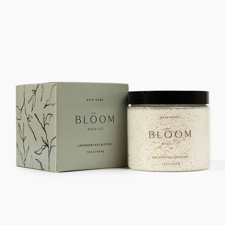 Clear glass jar with black lid and black text saying, "Bloom Bath Co. Eucalyptus Lavender Bath Soak". Packaged in a square green box with outline images of plant leaves.