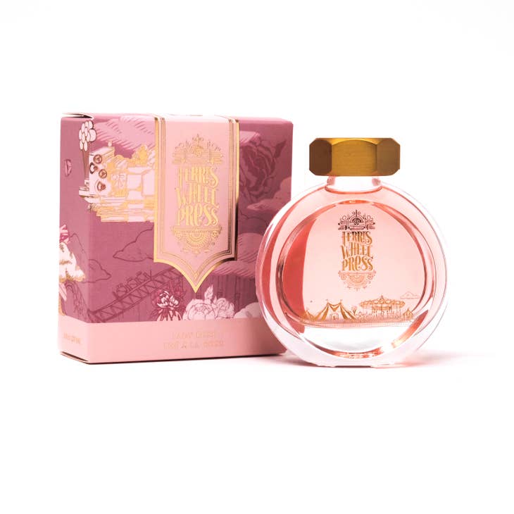 Round glass bottle with gold cover and gold text saying, "Ferris Wheel Press" with images of a carnival on front of bottle. Ink is pink. Packaged in square pink box with images of rose flowers.