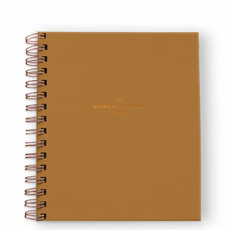 Notebook with orange mustard cover with gold foil text saying, “Quarterly Overview Planner”. Gold coil binding on left side.