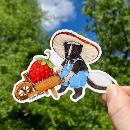 Sticker in the image of a skunk wearing blue overalls and hat pushing a red wheel barrow holding a red strawberry inside.