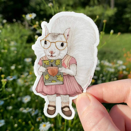 Sticker in the image of a white squirrel wearing a pink dress, reading glasses, pink socks and brown shoes holding a book about acorns.