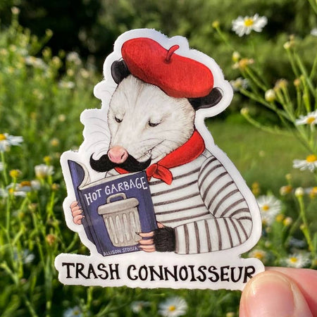 Sticker in the image of an opossum dressed in a white striped shirt, red beret, red scarf and a black mustache reading a book about hot garbage. Black text saying, “Trash Connoisseur”.