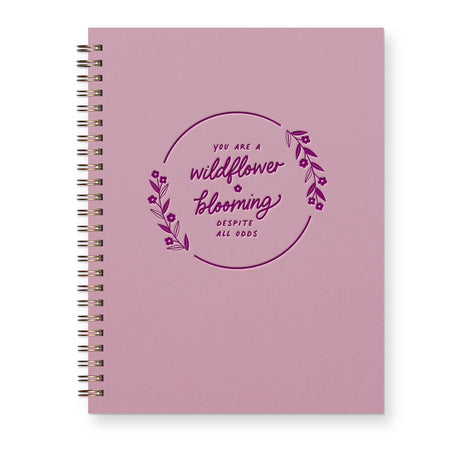 Light purple cover with purple text saying, “You Are A Wildflower Blooming Despite All Odds”. Image pf a floral wreath surrounding text. Metal spiral coil binding on left side.
