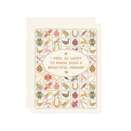 Ivory card with tan text saying, “I Feel So Lucky to Know Such a Beautiful Person”. Image of a patterned quilt with pictures of a ladybug, goose, horseshoe, rainbow, flower, wishbone, cat, rabbit and four leaf clover inside of quit. A cream envelope is included.