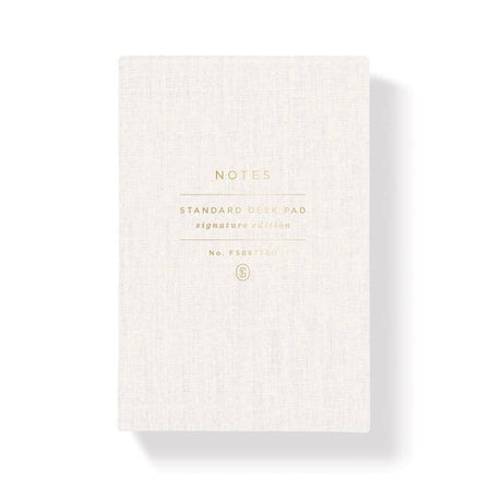 White linen notebook with gold foil text saying, “NTOES Standard Desk Pad Signature Edition”. 