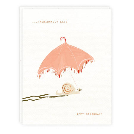 Ivory card with gold foil text saying, “Fashionably Late Happy Birthday.” Image of a tan snail holding a pink umbrella. A white envelope is included.