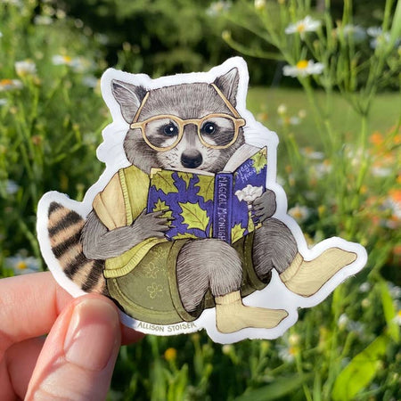 Sticker in the image of a raccoon wearing a green shirt and green shorts, reading glasses and yellow socks holding a book about flowers.