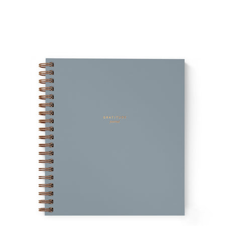Notebook with steel blue cover with gold foil text saying, “Gratitude Journal”. Gold coil binding on left side.