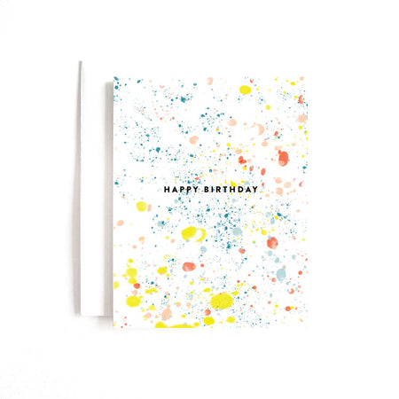 White card with black text saying, “Happy Birthday”. Images of colored paint splatters across card. A white envelope is included.