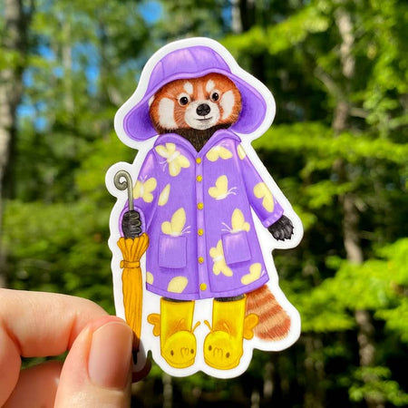Sticker in the image of a red panda wearing a purple rain jacket and yellow rain boots holding a yellow umbrella.