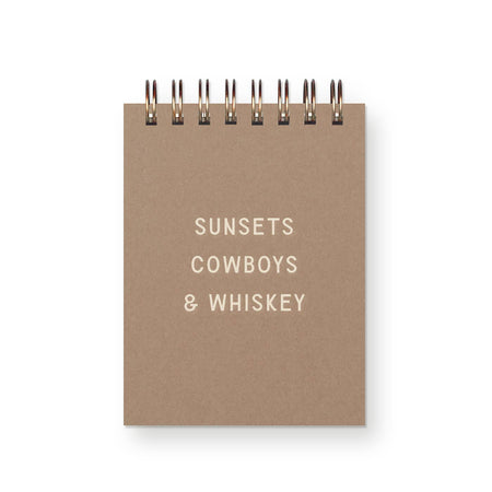 Brown craft paper cover with white text saying, “Sunsets Cowboys & Whiskey”. Metal spiral coil binding across the top.