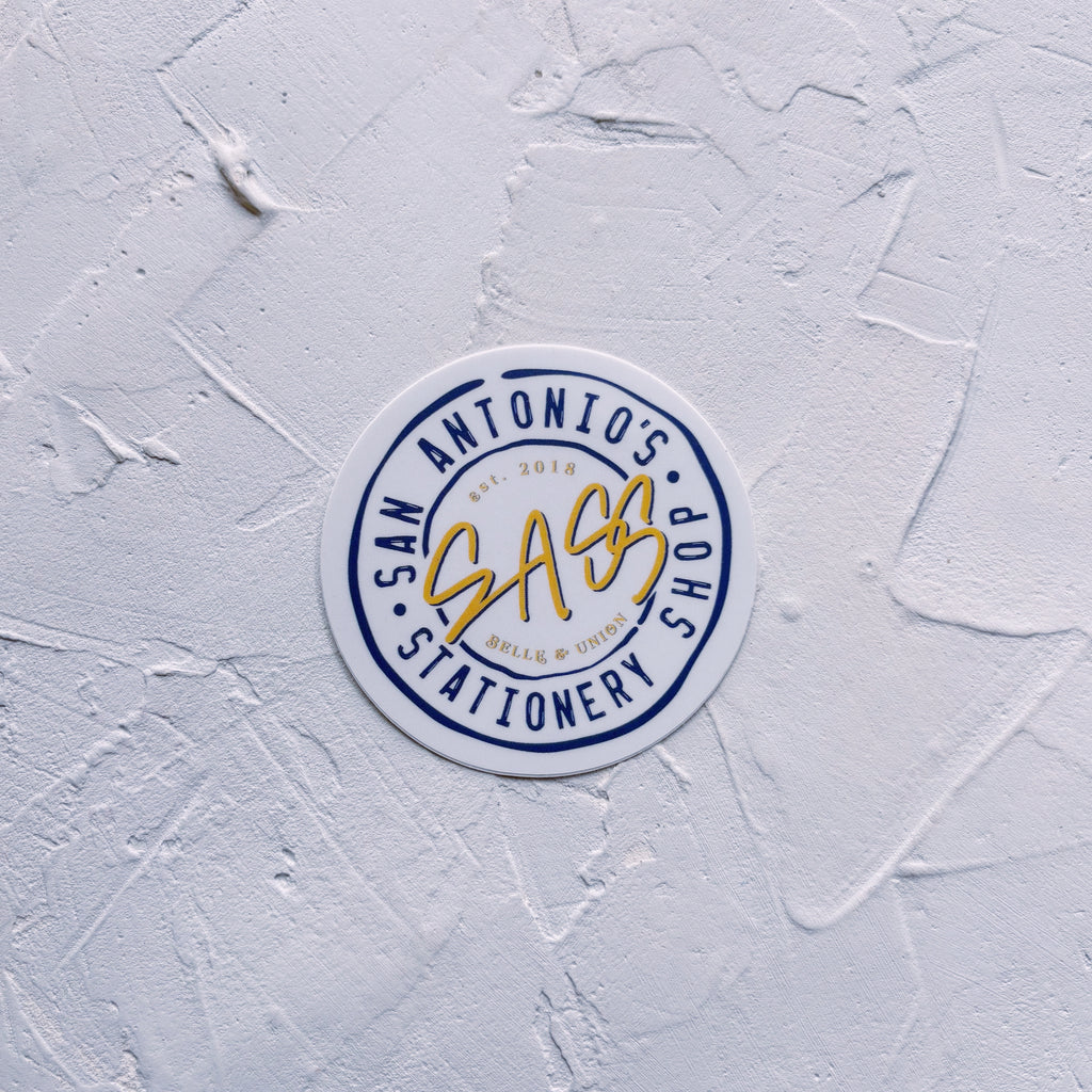 Round Belle & Union sticker with navy text and "SASS" in yellow text in the middle. Sticker is on textured background.
