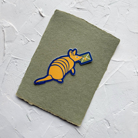 Sticker of yellow armadillo with a green envelope in its mouth, bordered with navy.  Sticker is on green textured paper. 