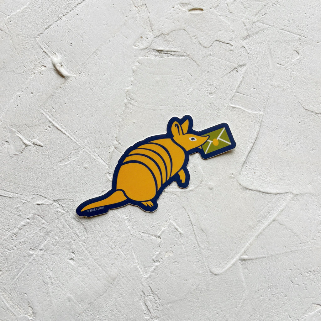 Sticker of yellow armadillo with a green envelope in its mouth, bordered with navy.  Sticker is on white textured background.