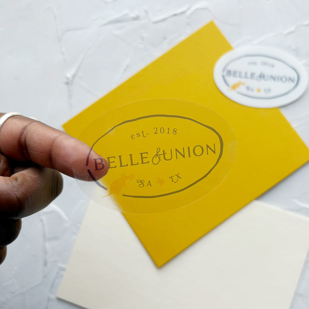 a finger displaying an oval belle & union sticker with navy text and yellow Texas and armadillo icon. Background features the same sticker, overlayed on yellow paper
