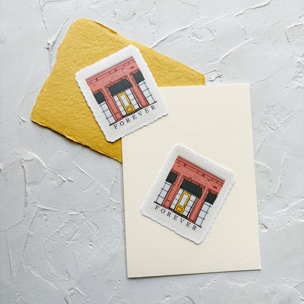 Two Belle & Union branded stickers of brick red building and yellow door. Stickers on yellow and cream paper, on white textured background.