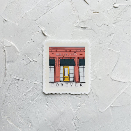 Belle & Union branded sticker of brick red building and yellow door. Sticker on white textured background.