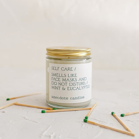 Glass candle with gold lid on textured background, with green matchsticks
