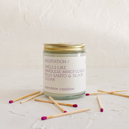 Glass candle with gold lid on textured background, with purple matchsticks