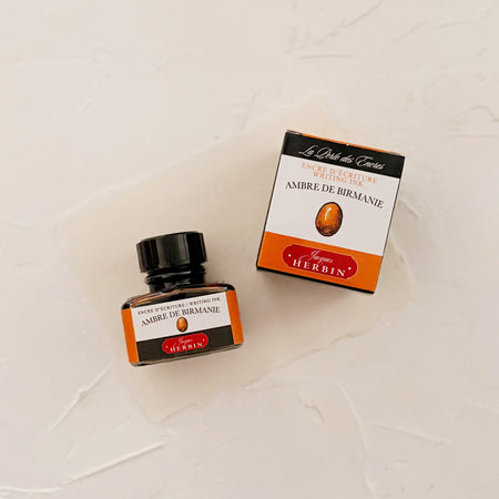 Glass square bottle with black cap. White label with black text saying, “Ambre De Birmanie” and white text saying, “HERBIN”. Orange stripe on each side of bottle - on textured background