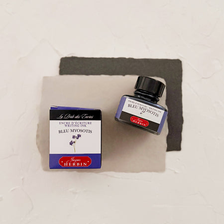 Glass square bottle with black cap. White label with black text saying, “Bleu Myosotis” and white text saying, “HERBIN”. Lavender stripe on each side of bottle - next to a box with the same label. both on textured background