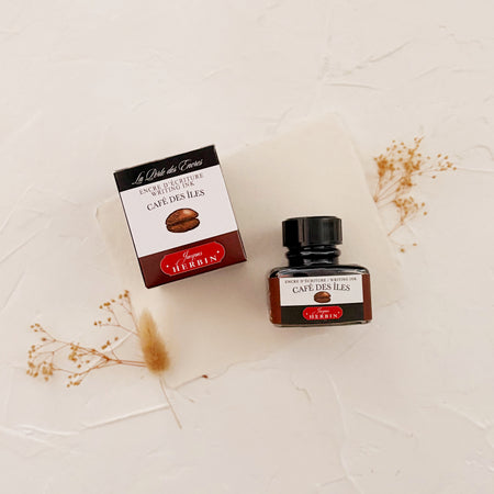 Glass square bottle with black cap. White label with black text saying, “Café Des Iles” and white text saying, “HERBIN”. Brown stripe on each side of bottle. bottle is next to a box with the same label. both on textured background and cream dried florals 