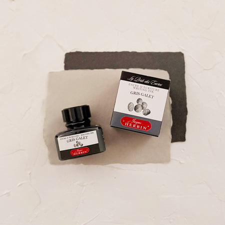 Glass square bottle with black cap. White label with black text saying, “Gris Galet” and white text saying, “HERBIN”. Gray stripe on each side of bottle. bottle is next to a box with the same label. both on textured gray background
