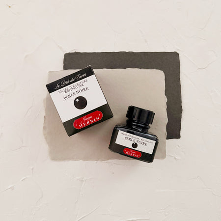 Glass square bottle with black cap. White label with black text saying, “Perle Noire” and white text saying, “HERBIN”. Black stripe on each side of bottle. bottle is next to a box with the same label. both on textured gray background