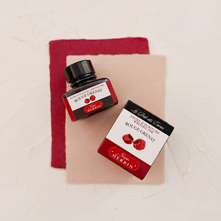 Glass square bottle with black cap. White label with black text saying, “Rouge Grenat” and white text saying, “HERBIN”. Red stripe on each side of bottle. bottle next to box with the same label. both on textured background