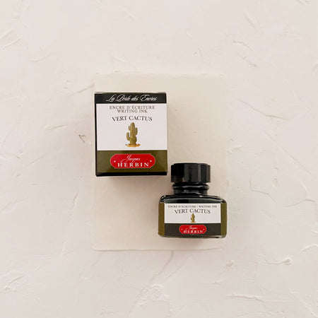 Glass square bottle with black cap. White label with black text saying, “Vert Cactus” and white text saying, “HERBIN”. Green stripe on each side of bottle. bottle next to box with same label. Both on textured background.